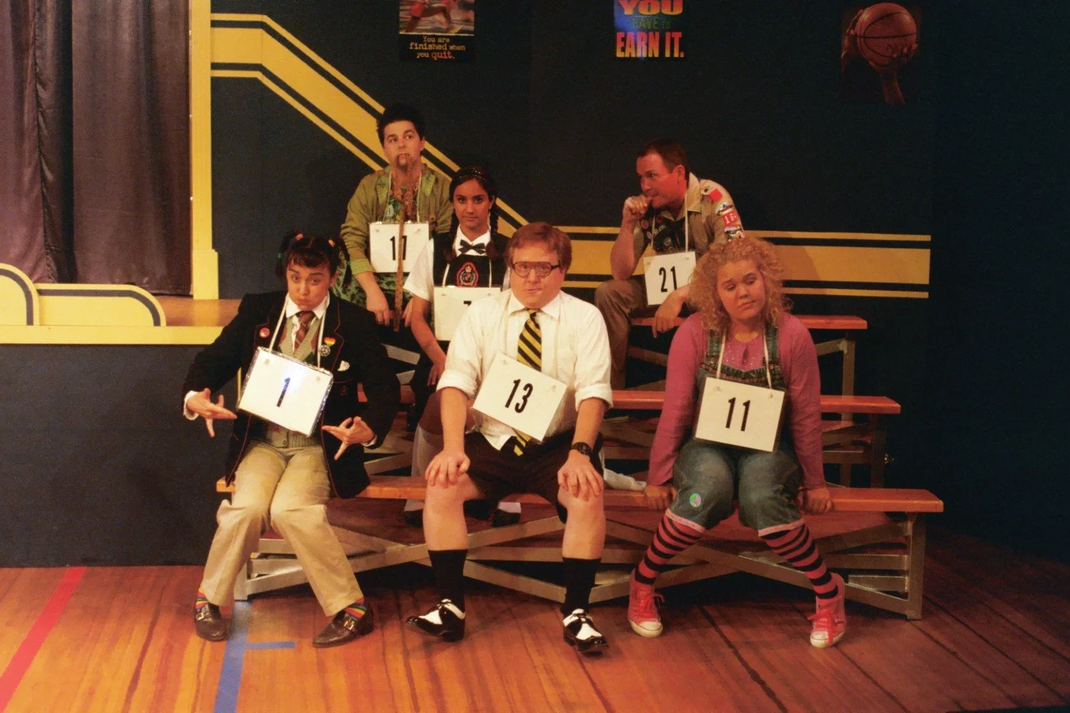 Group of people in costumes sitting on bleachers, each wearing numbered tags around their necks, suggesting a humorous or theatrical setting, with a backdrop featuring motivational posters.