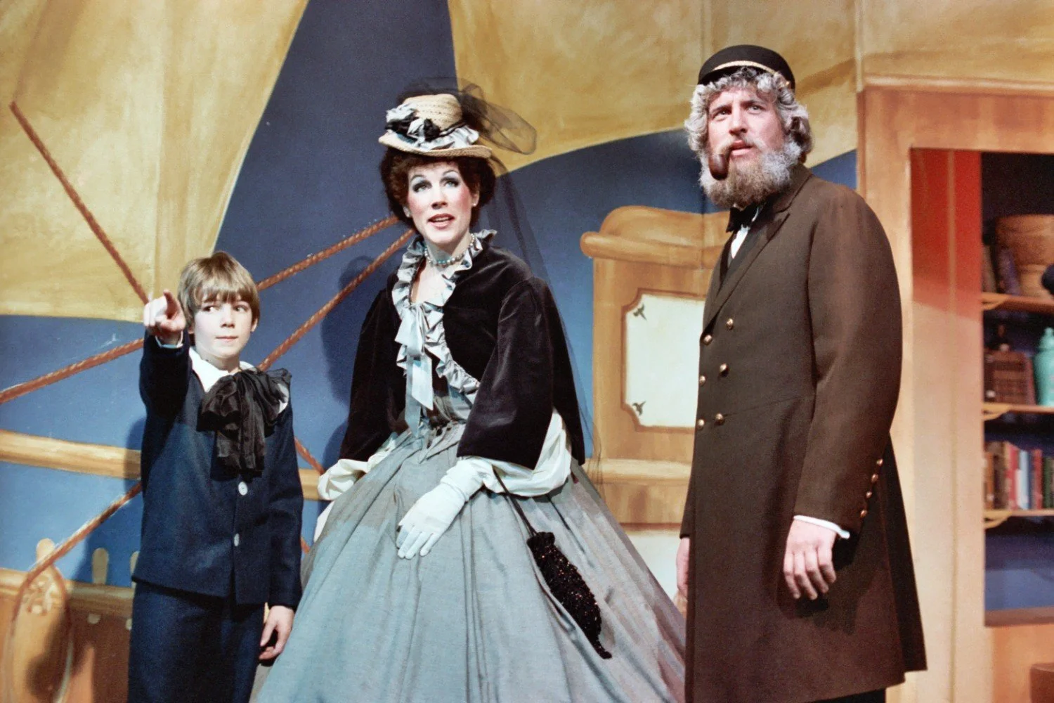 Scene from a theatrical play with a boy pointing, a woman in period dress, and a bearded man with a pipe.