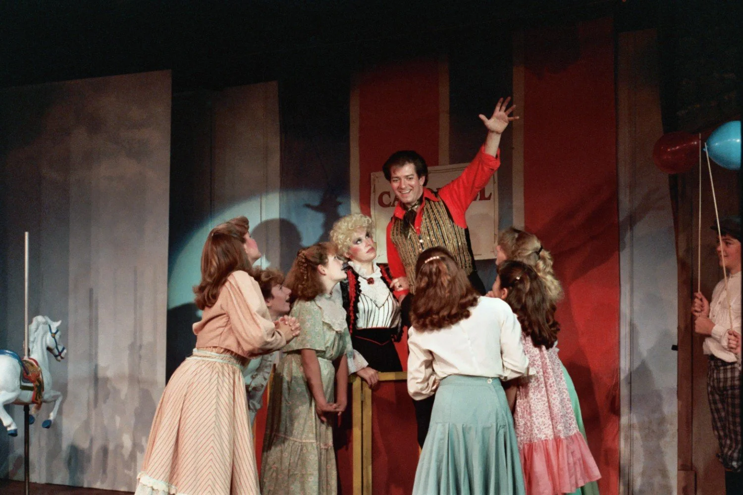 Group of people in period costumes on stage during a theatrical performance, with a man in a red shirt raising his arm and children gathered around.