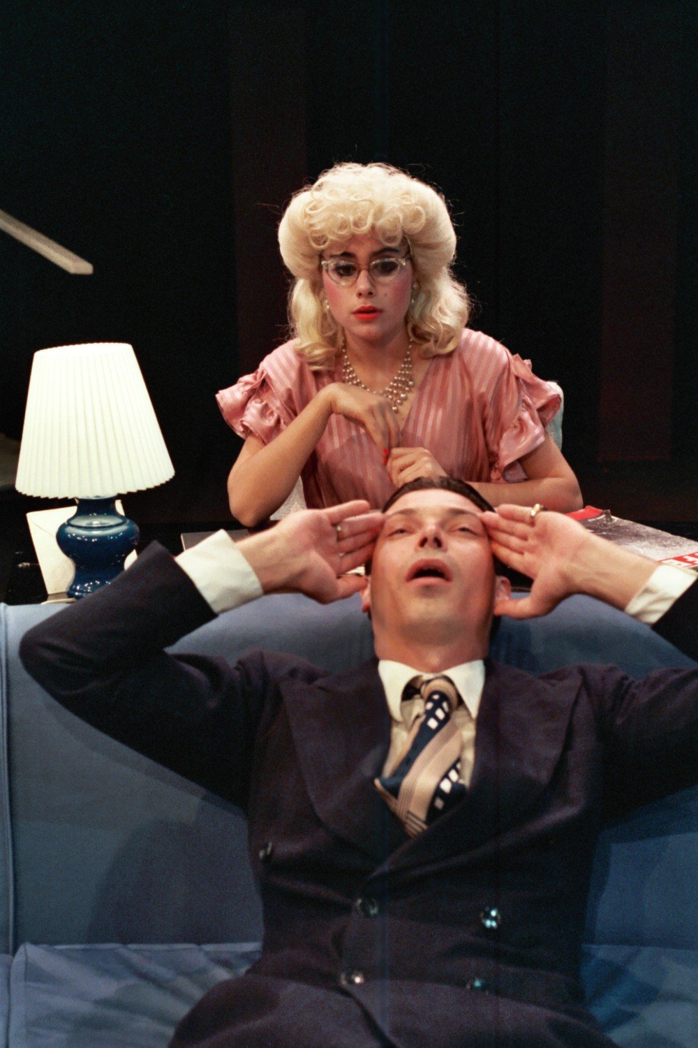 A scene from a theatrical performance showing a woman with blonde hair and glasses seated behind a man in a suit reclining on a blue couch with his hands on his head. A small table lamp is visible next to the woman.