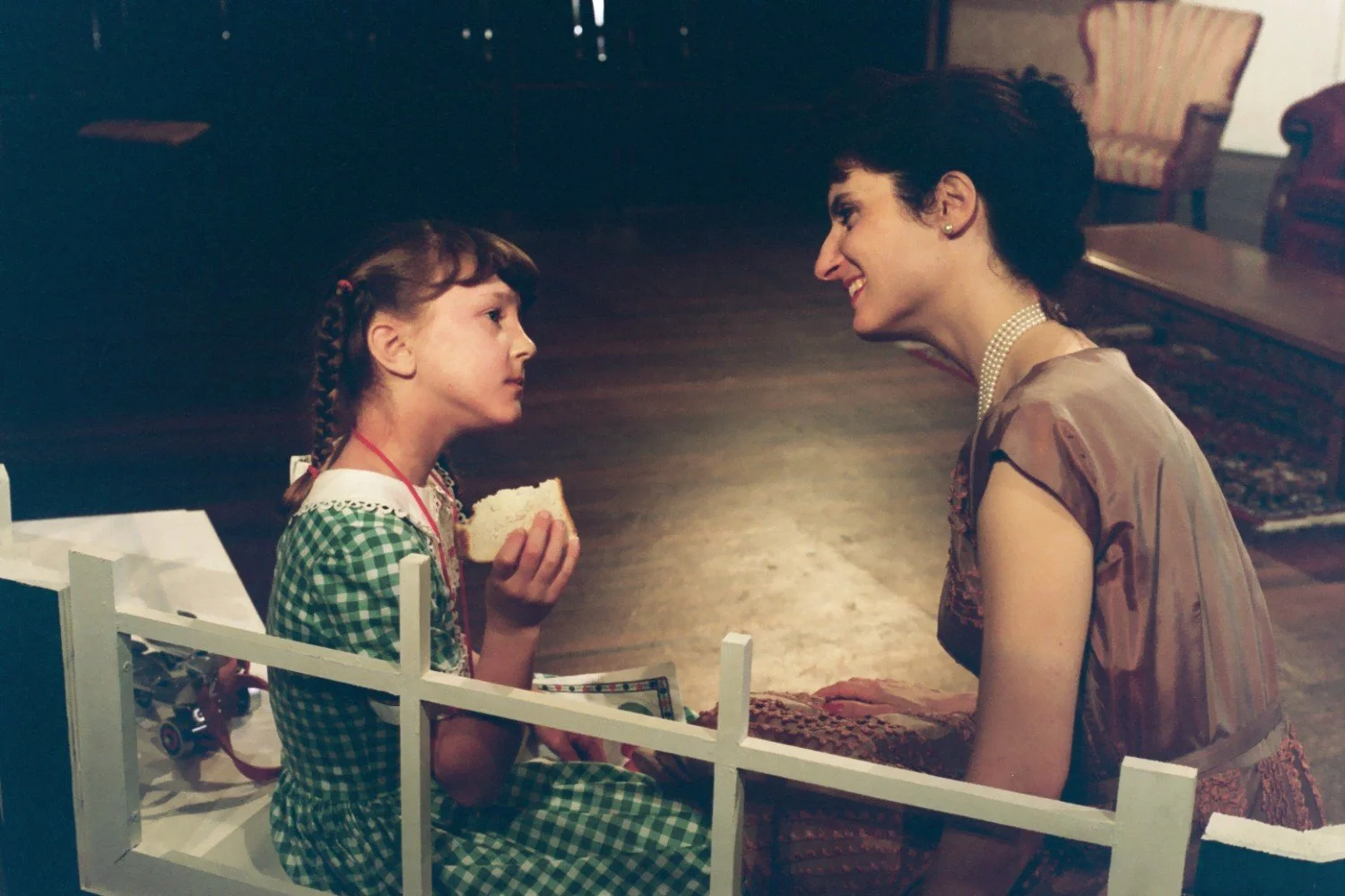 A young girl in a green checkered dress sits and holds a piece of bread, looking intently at a woman kneeling in front of her. The woman, wearing a pearl necklace, listens and smiles warmly at the girl. They are seated indoors, with furniture visible