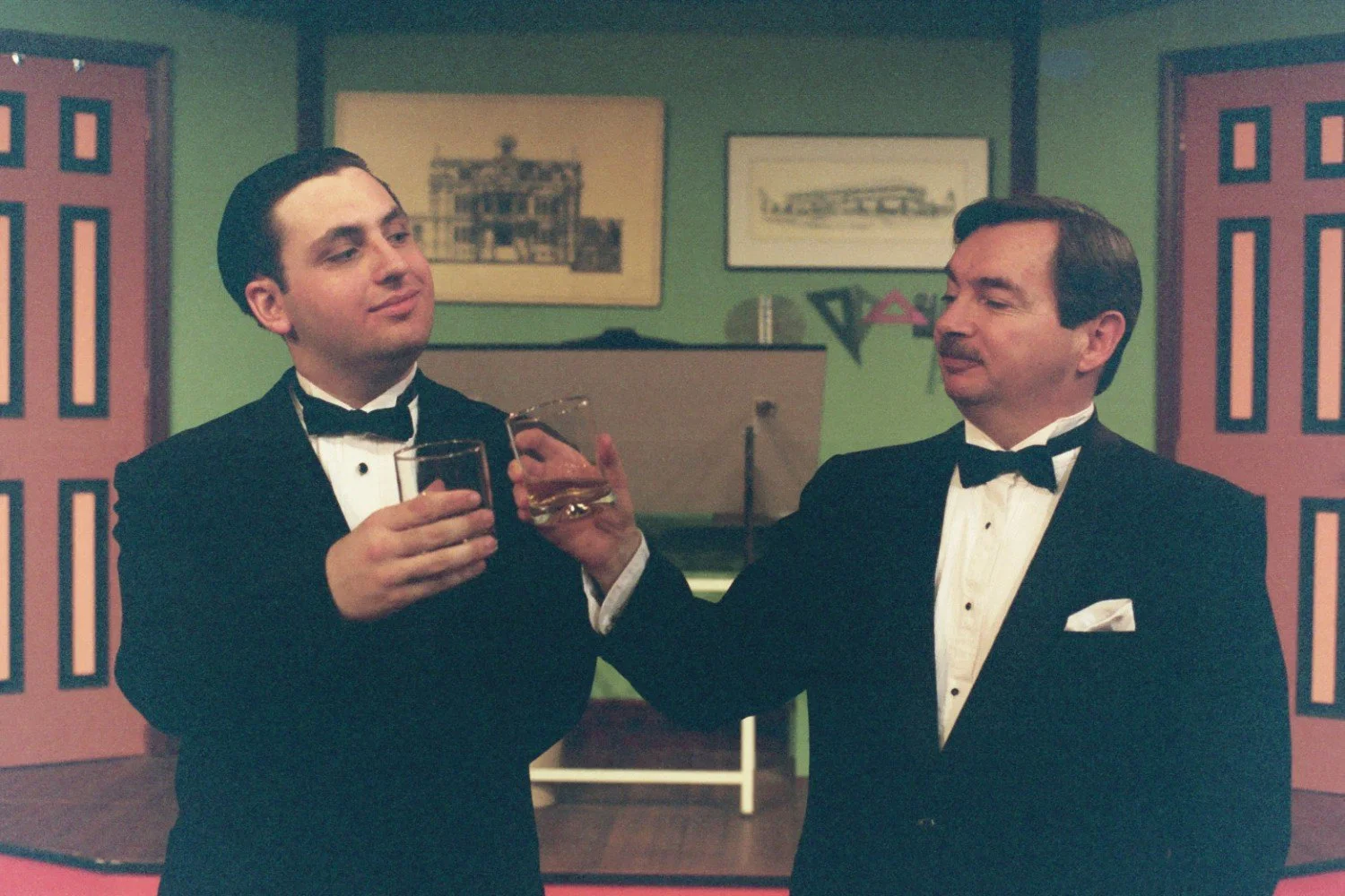 Two men in tuxedos clinking glasses in a room with framed pictures on the wall and two doors in the background.