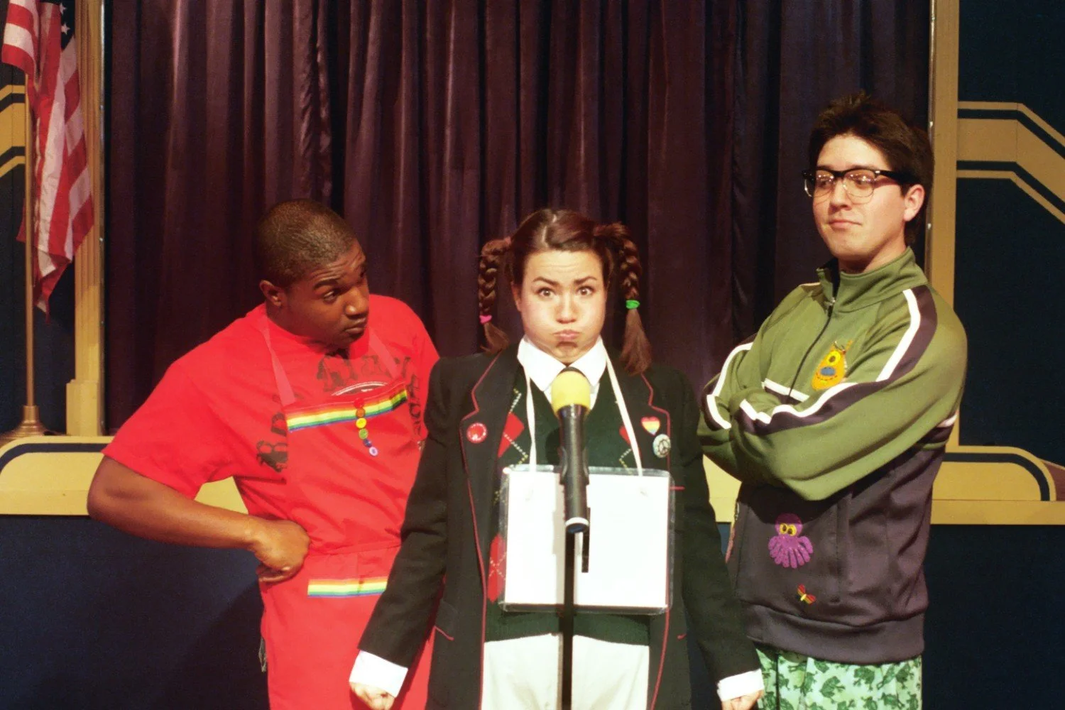 Three people on stage, one woman in a school uniform with pigtails stands at a microphone, flanked by two men wearing casual clothing.