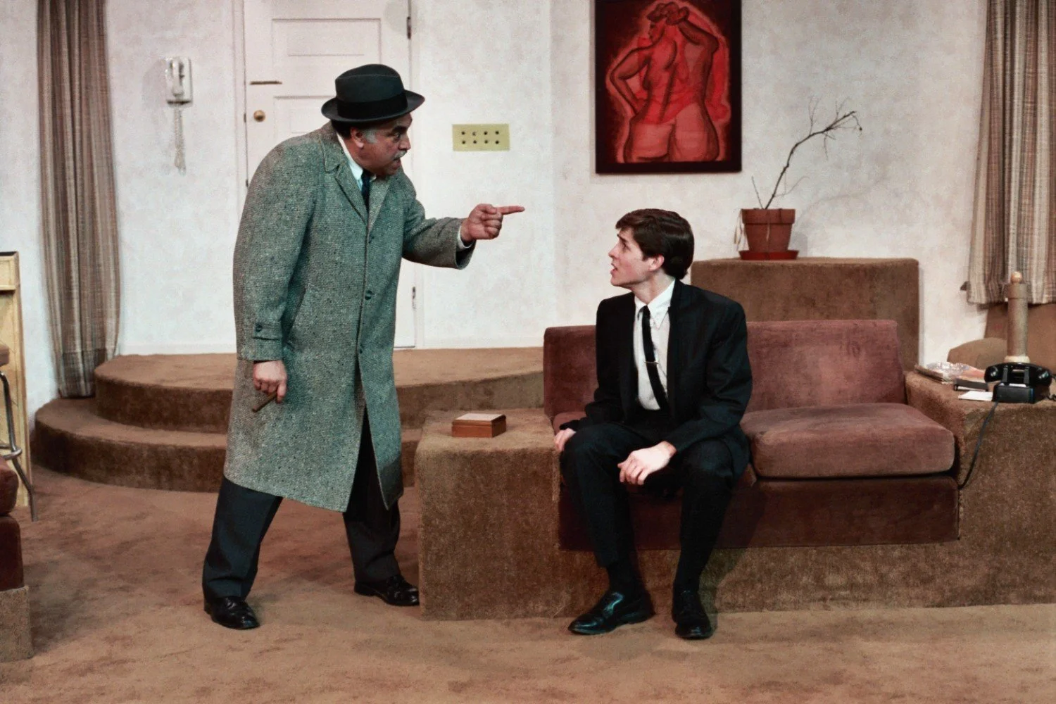 Two men in a room; one standing and pointing, the other sitting and looking up at him, with a couch, retro phone, and artwork on the wall.