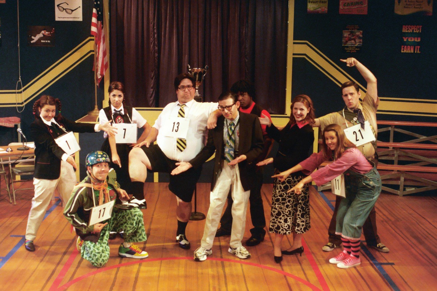 A group of people in costumes posing indoors, with some wearing numbers like a spelling bee or competition. They are standing on a gymnasium floor, with a trophy and an American flag in the background. The scene resembles a comedic or theatrical even