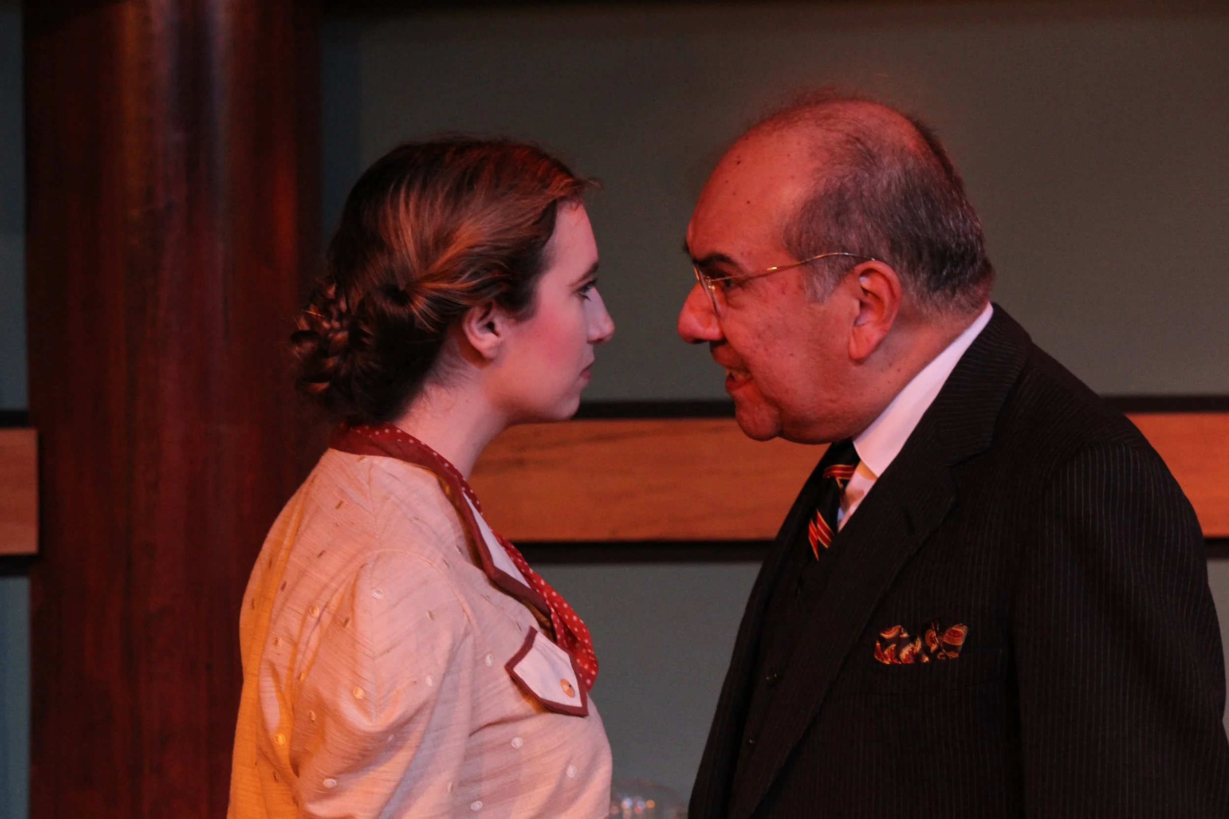 Two actors in a theatrical scene, a woman and a man, facing each other closely. The woman has a braided hairstyle and is wearing a vintage dress, while the man is in a suit and tie with eyeglasses. The background features wooden paneling.