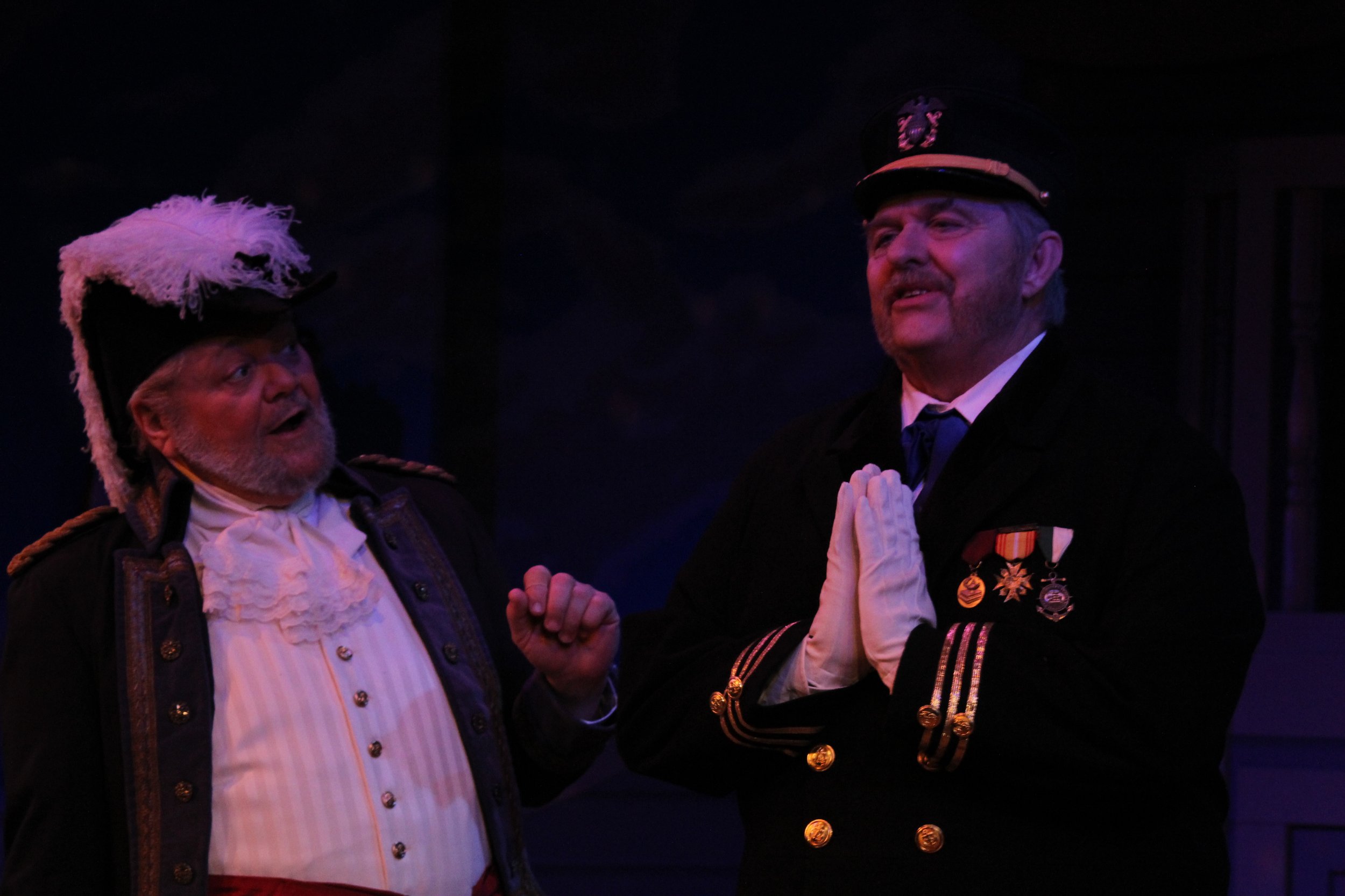 Two men dressed in historical military costumes, one with a feathered hat and the other with medals and gloves, on stage performing a theatrical scene.