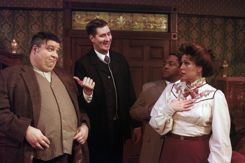 Four people dressed in vintage clothing are in a room with ornate wallpaper and a wooden door. The man on the far left is wearing a checkered blazer and gesturing with his hand. The man next to him is wearing a suit and smiling. Another man looks on