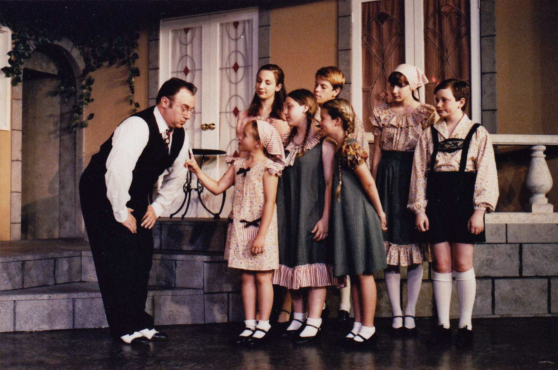 A group of people on stage in period costumes, part of a theatrical production. A man in a vest and tie leans toward a line of children, who are dressed in old-fashioned clothing, including dresses, aprons, and suspenders. They are standing on a set