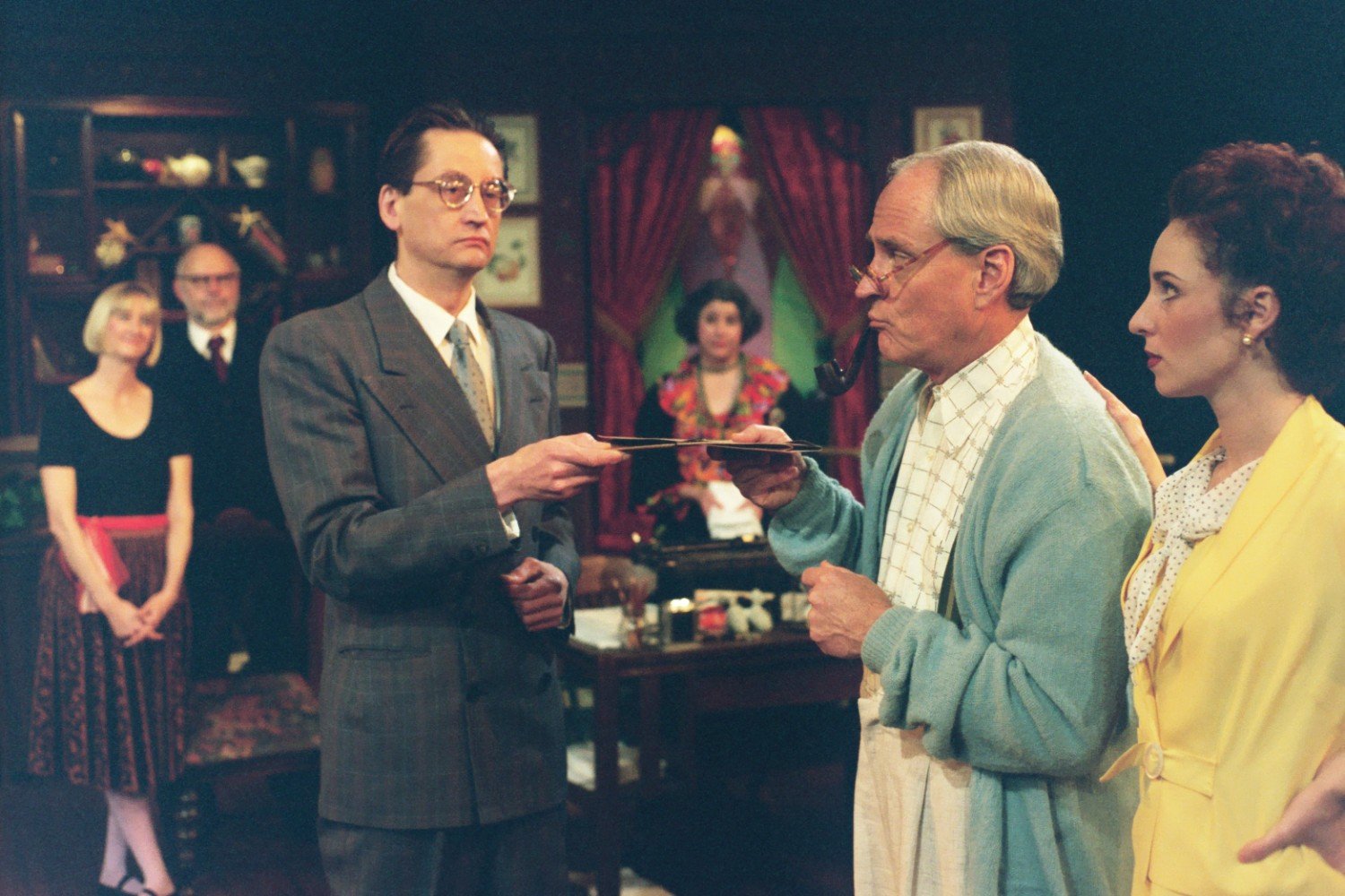 Group of actors on stage performing a scene in a traditional setting with vintage costumes, including suits and dresses. One man holds a tray, another smokes a pipe. Bookshelves and curtains in the background.