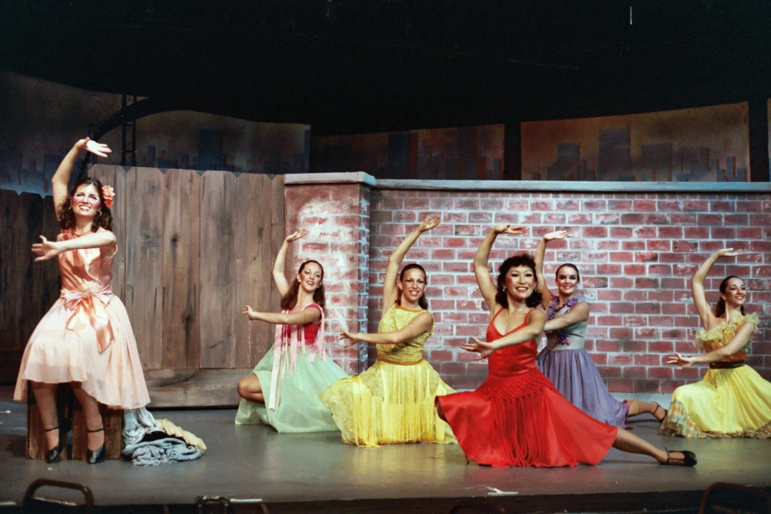 A group of dancers performing on stage in colorful dresses against a wooden and brick backdrop.