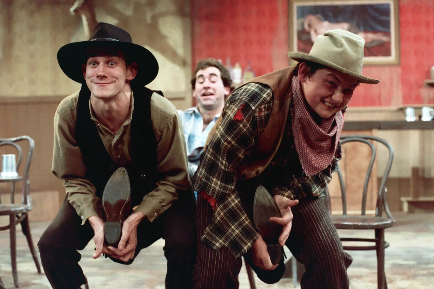 Three people in a comedic theater scene dressed as cowboys, with two holding shoes and smiling, in a Western-style setting.