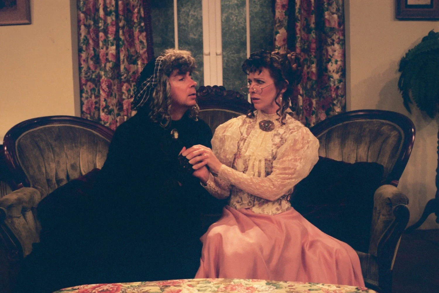 Two people in period costumes sitting on a vintage sofa, engaged in an emotional conversation, with floral curtains in the background.