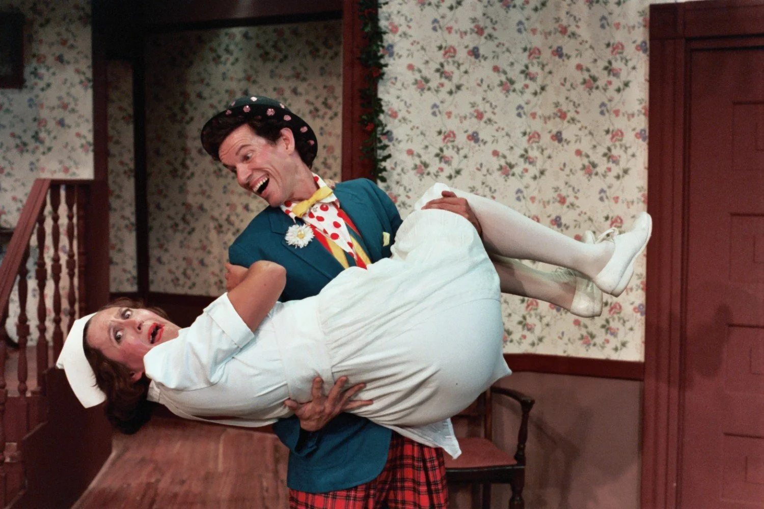 A person in a clown outfit holding another person dressed as a nurse in a humorous, dramatic pose. The background features a floral wallpaper and wooden interior.