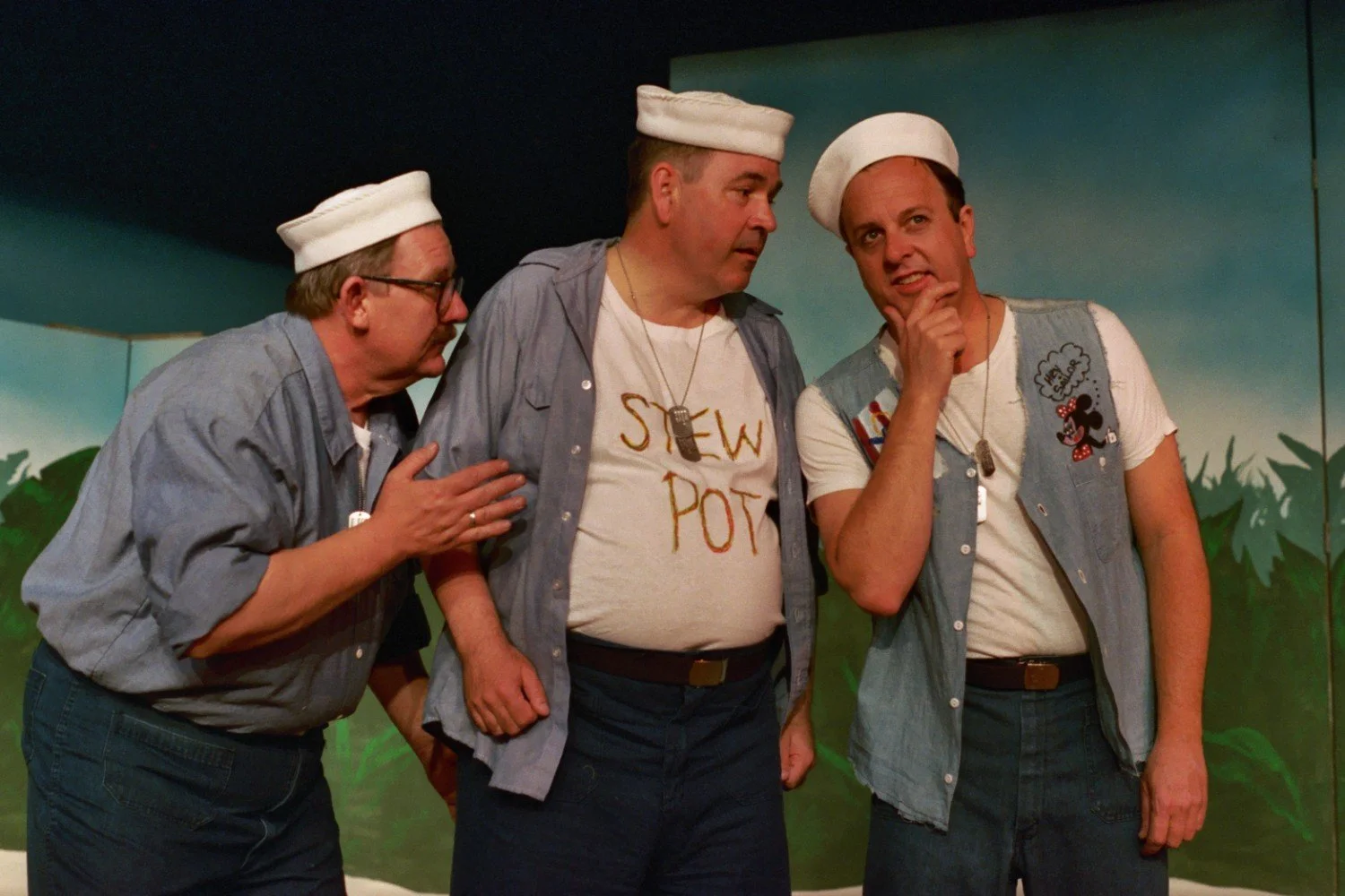 Three men dressed as sailors in a theatrical scene, wearing white caps and denim shirts, engaged in a conversation on stage with a painted backdrop.