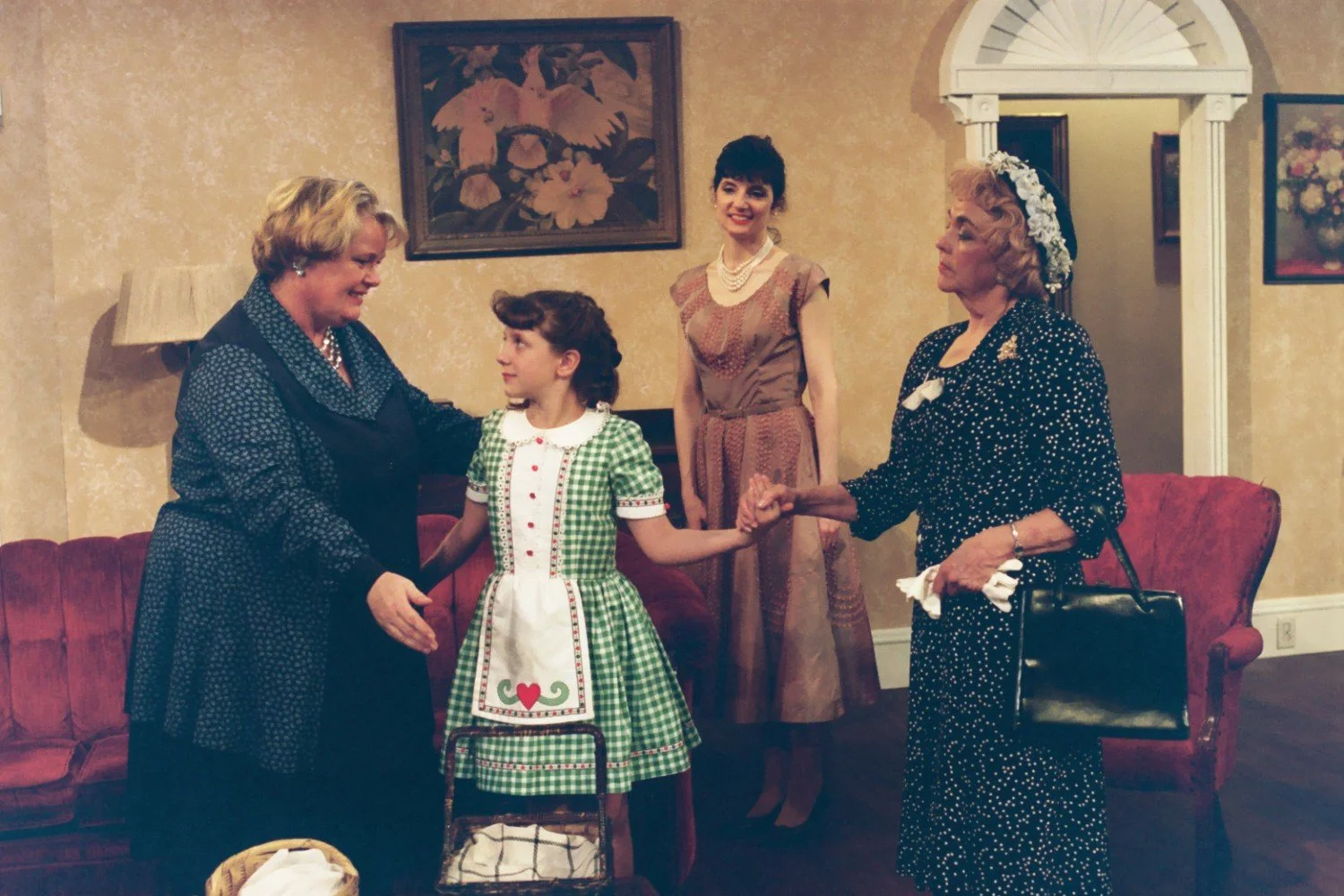 Scene from a play with four people in period costumes, two women holding hands with a girl in a green checkered dress, another woman stands smiling in the background.