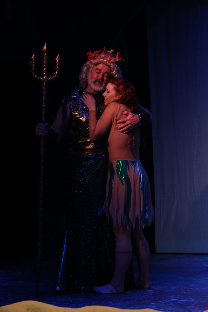Two performers in a theatrical scene, one wearing a crown and holding a trident, comforting the other on stage.