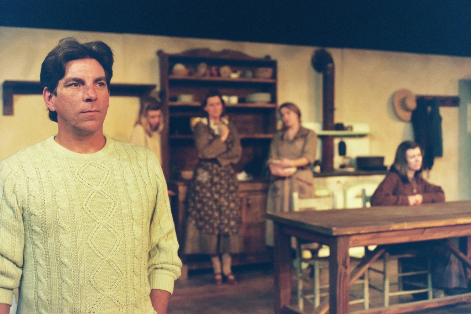 A man in a knitted sweater stands in the foreground of a theater stage set, with four women in period costumes in the background, suggesting a historical drama setting in a rustic kitchen or dining room.