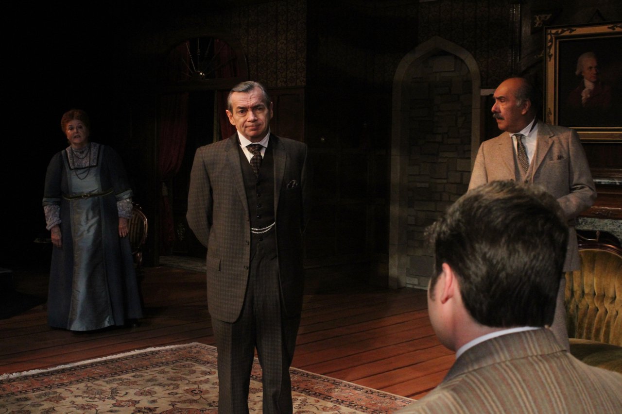 Four actors on stage in a period drama setting, with vintage furniture and decor. One male actor stands facing the audience, dressed in a dark suit. Another man stands to his right, while a woman in a long dress stands in the background. A fourth act