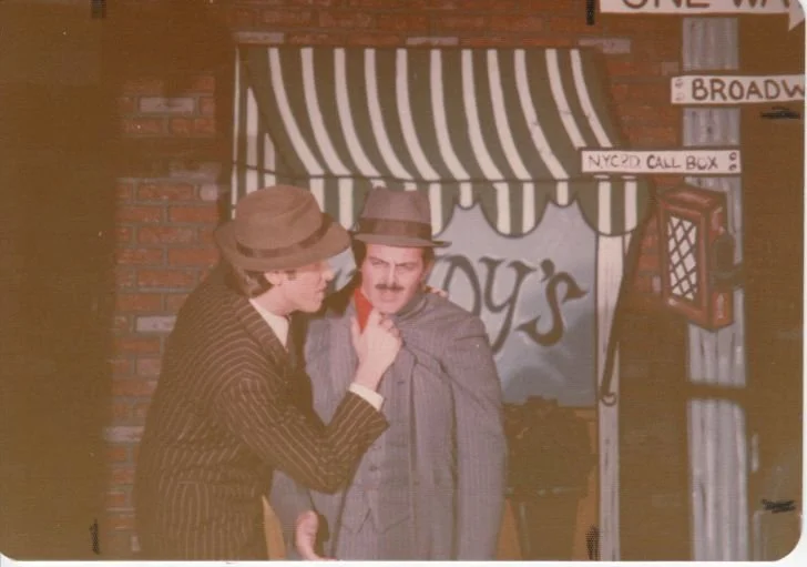 Two men wearing vintage-style suits and hats, with one man holding the other by his coat collar in a dramatic pose. They are standing outside a storefront with a striped awning and signs indicating "NYCPD Call Box" and "Broadway."