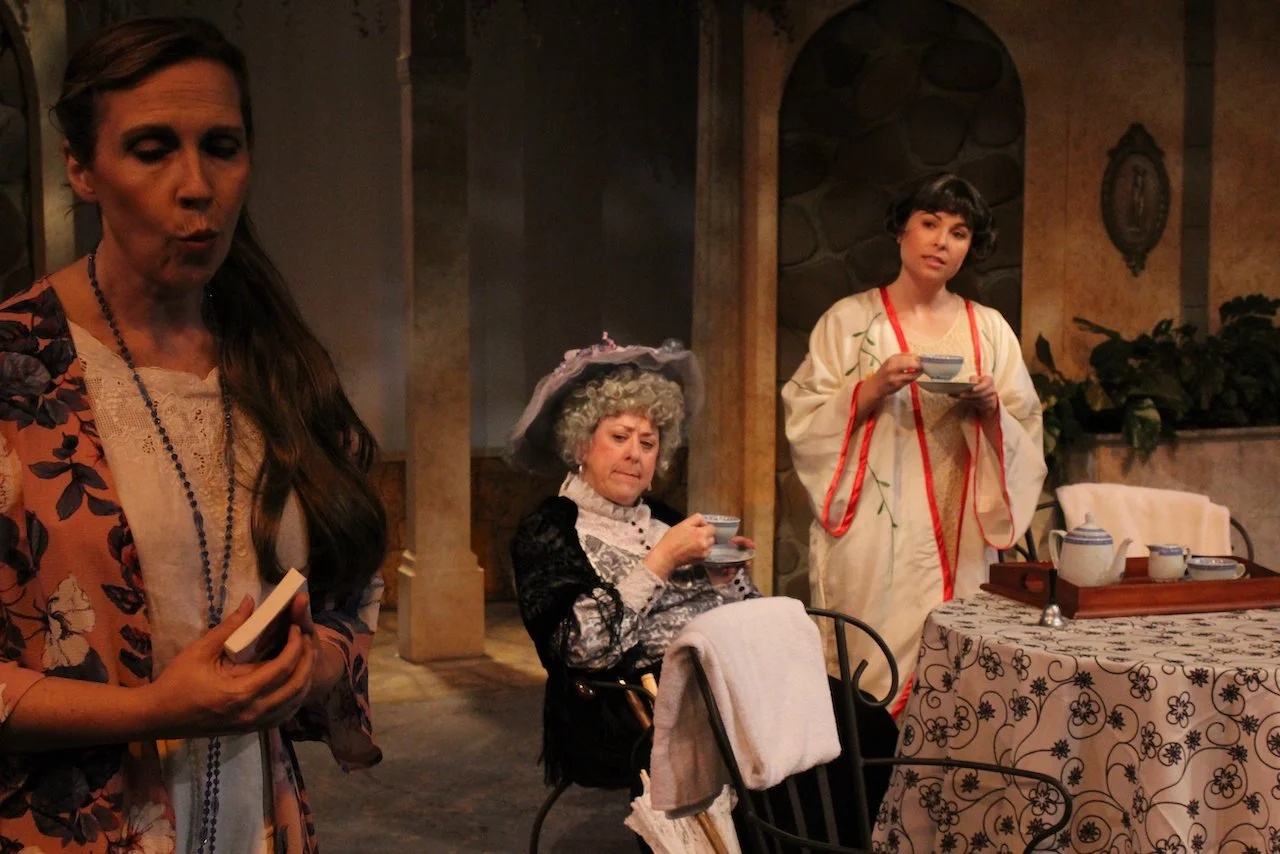 Three women in period costumes interacting in a theatrical setting, with a table set for tea, featuring teapots and a tray.