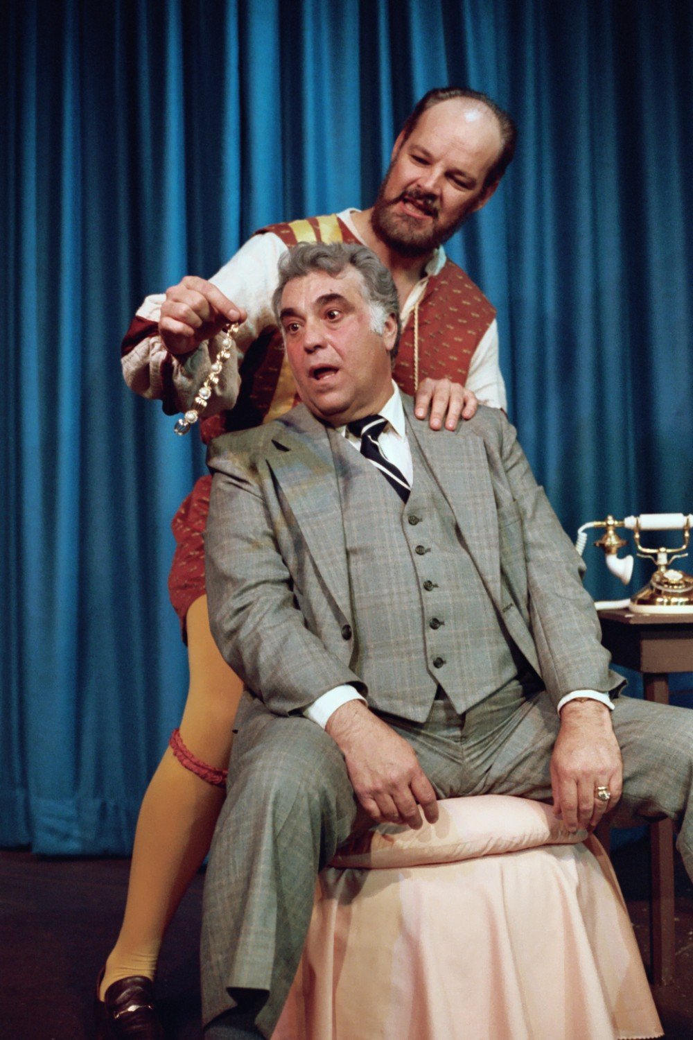 Two actors on stage in a comedic play; one seated in a gray suit and tie, the other standing behind in colorful costume holding jewelry; both with animated expressions; blue curtain backdrop.