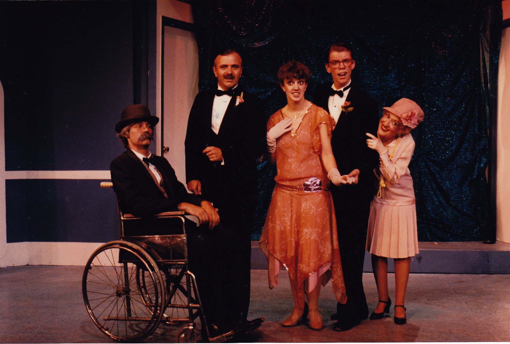 A theatrical scene with five people in vintage 1920s attire. A man in a wheelchair is on the left, wearing a hat. Two men in tuxedos stand in the center; one woman in a flapper dress is next to them. Another woman in a vintage hat and dress stands to