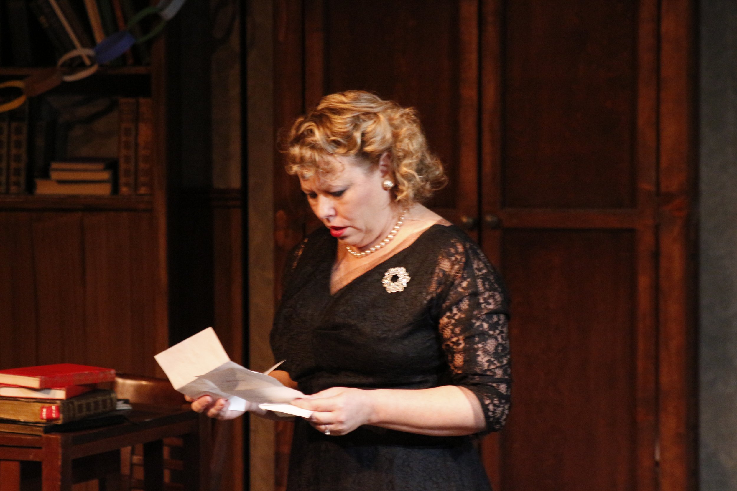 A woman in a black lace dress reads a letter, standing next to a wooden table with books, in a room with wooden furniture.
