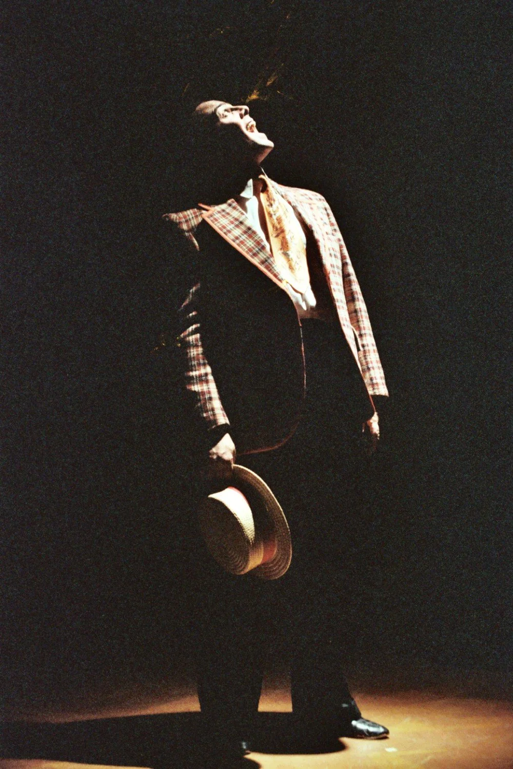 Person in checkered suit with straw hat singing on stage in dramatic lighting.