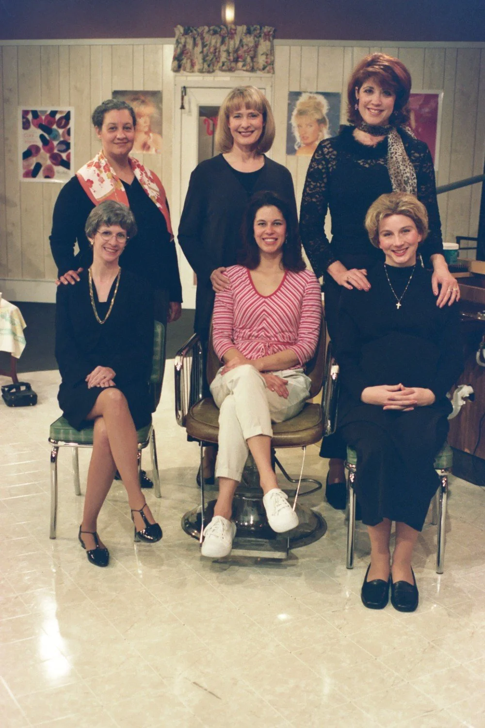 Group of six women posing in a hair salon setting, with posters on the walls and styled hair as a central theme.