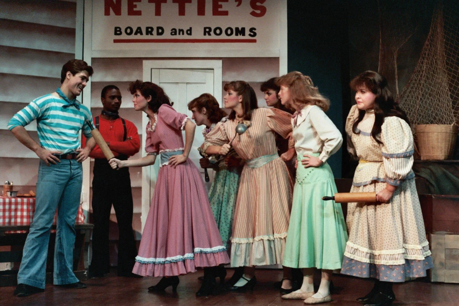A stage performance featuring a group of actors in period costumes in front of a set labeled 'Nettie's Board and Rooms.' A man in a striped shirt stands to the left, while several women in long dresses appear to confront him. Some women hold kitchen 