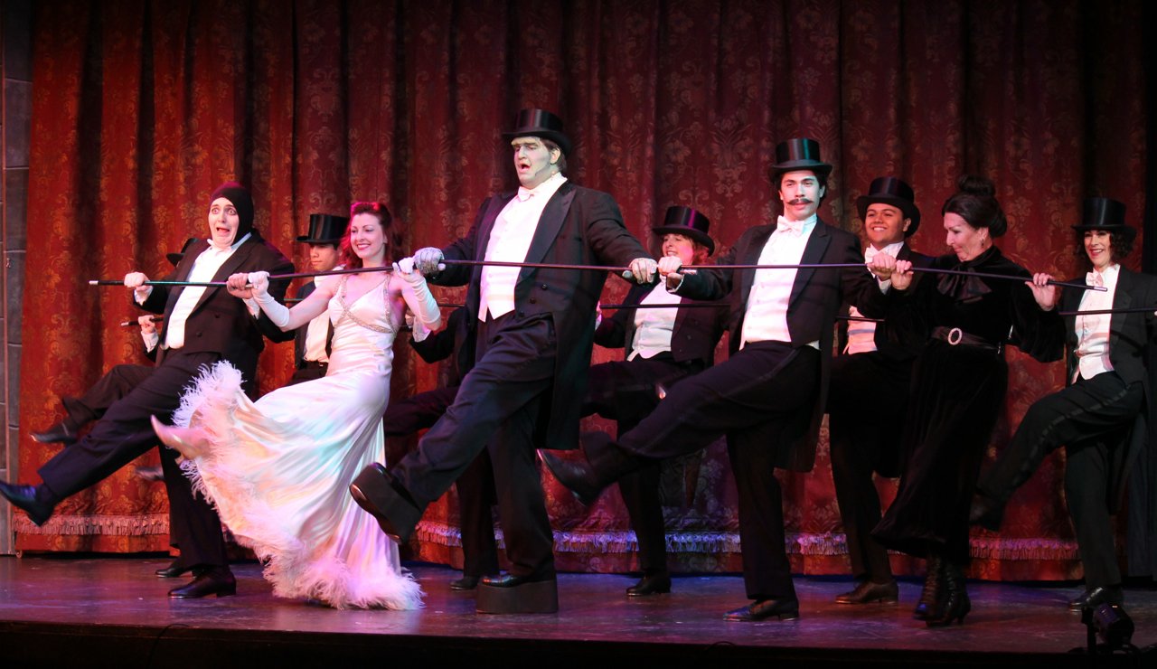 Theatrical performance with cast in formal attire, including top hats and gowns, dancing with a cane on stage.