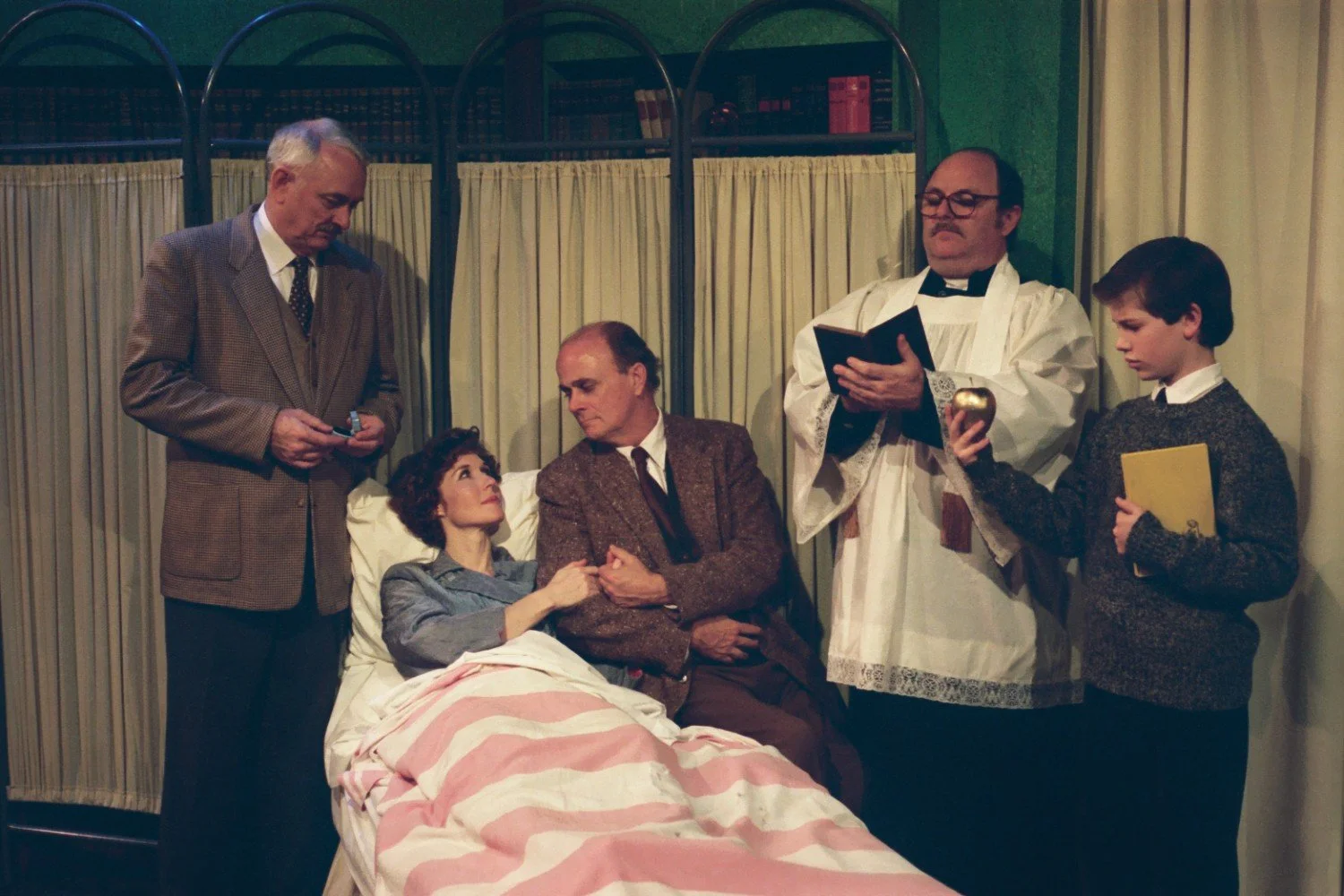 A woman in a hospital bed surrounded by four people, including a man in a priest outfit, holding a book and a young boy holding a golden cup. Another man is sitting on the bed holding the woman's hand while accompanied by an older man standing nearby