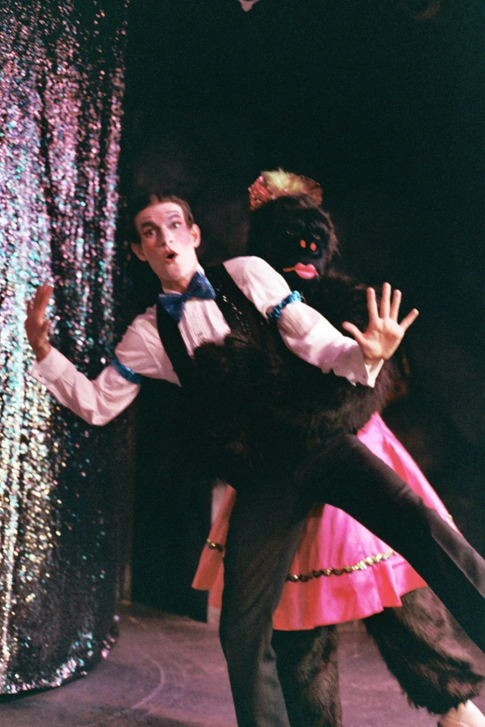 Performer in a white shirt and bow tie, with a person in a gorilla costume behind him, acting surprised on stage with a sparkling curtain backdrop.