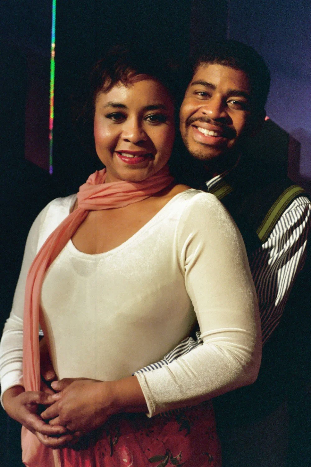 A smiling couple posing together in formal clothing, with the woman in a white dress and pink scarf, and the man in a striped shirt and vest.