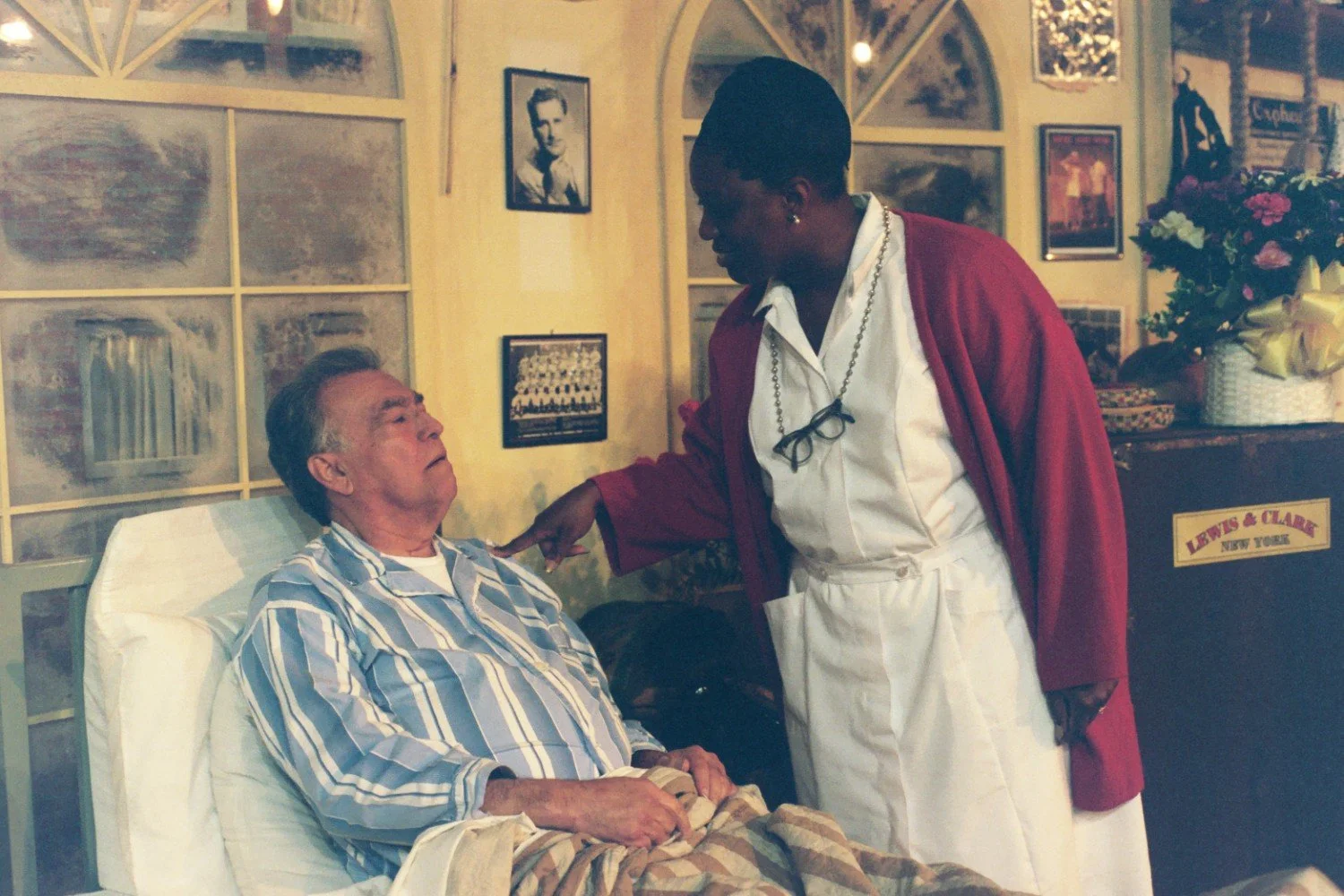 A nurse in a red cardigan speaking to a man in a hospital gown, seated in a hospital bed inside a room adorned with various framed photographs and decorations.