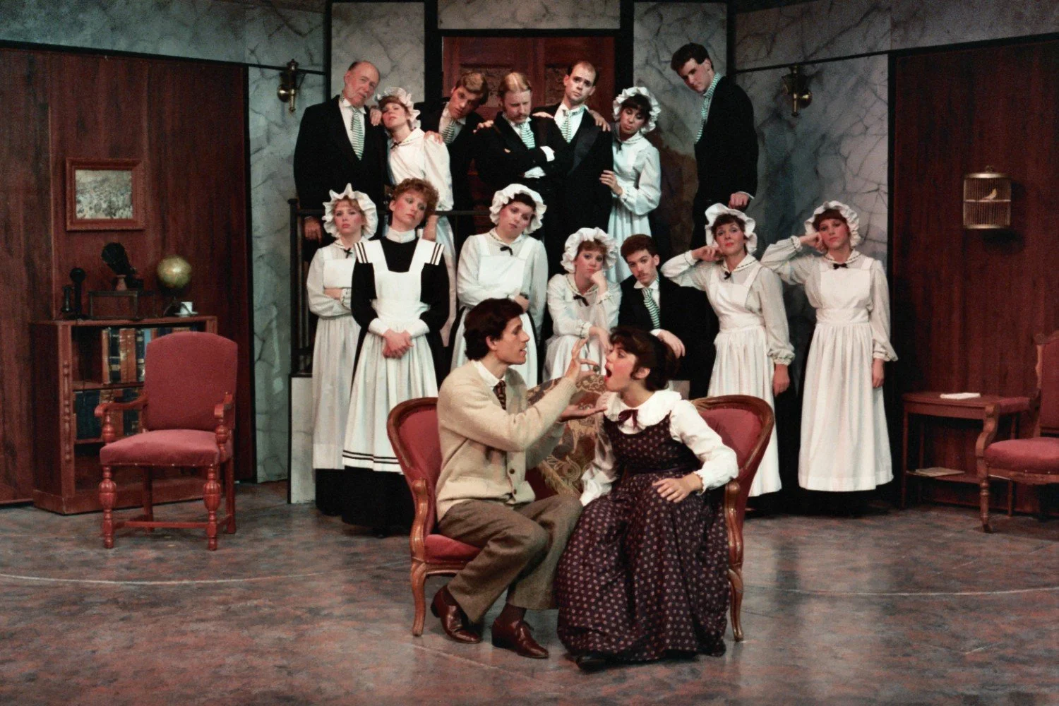 Theatrical stage scene with actors dressed in period costumes, including maids, butlers, and seated couple in Victorian-style garments, set in a classic interior with chairs and bookshelves.