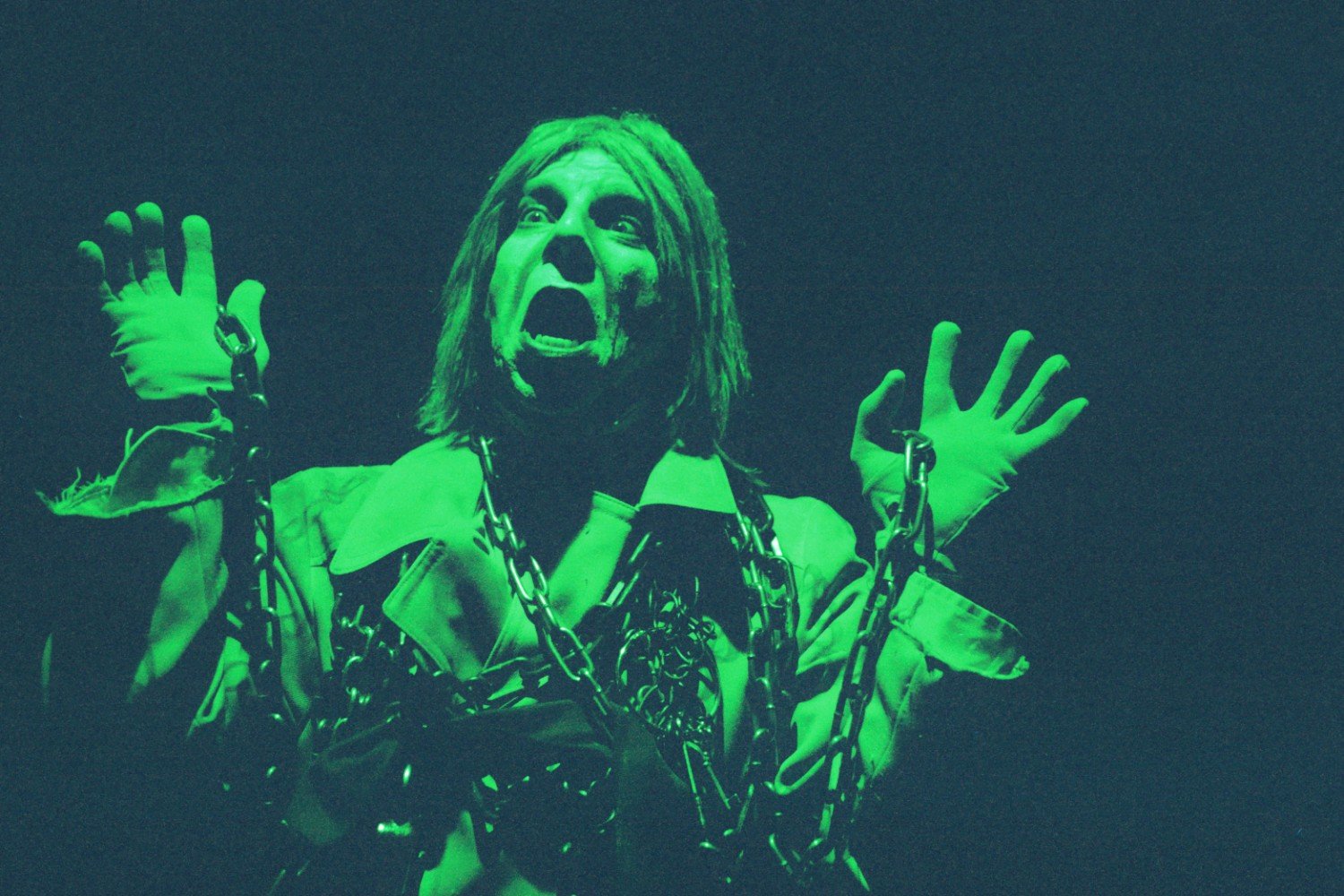 A person wearing theatrical makeup and costume, with chains draped over their shoulders, set against a dark background. The image has a greenish tint, creating a dramatic effect.