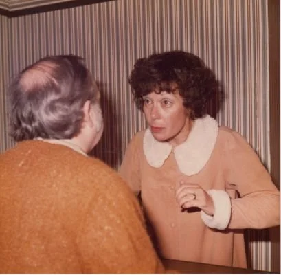 Two people inside a room with striped wallpaper, one with back to camera, other making an expressive gesture.