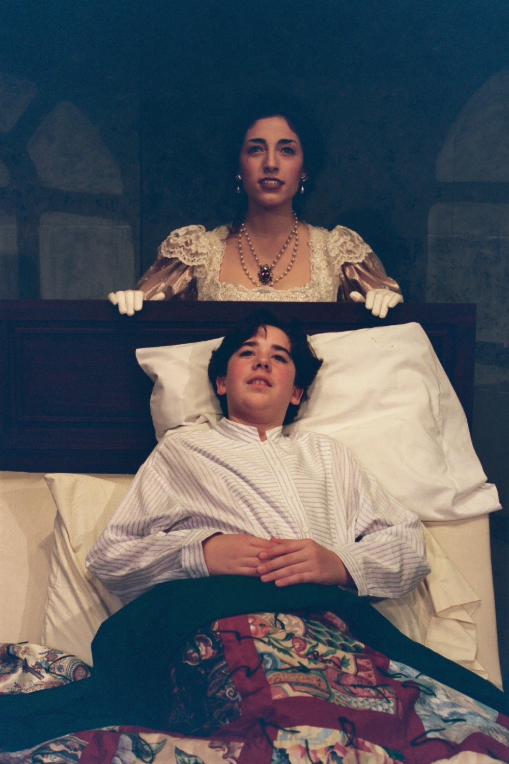 A person dressed in historical attire stands by a bed, while another person is lying in bed covered with a quilt. The standing person is wearing a lace dress and gloves, and the person in bed is in a striped nightshirt.