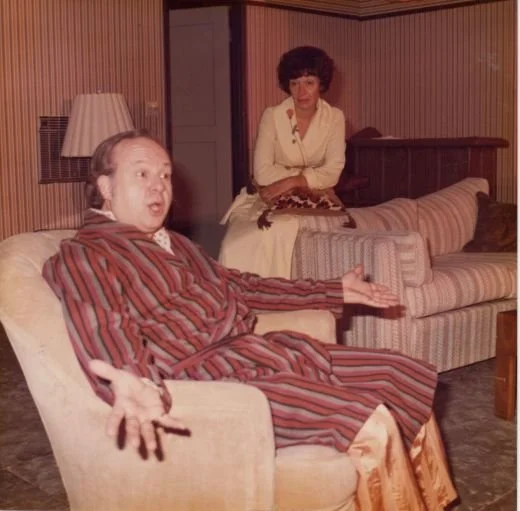 A man in a striped robe sits in a chair gesturing with his hands, while a woman in a white dress sits on a couch behind him. The room has striped wallpaper and a lamp.