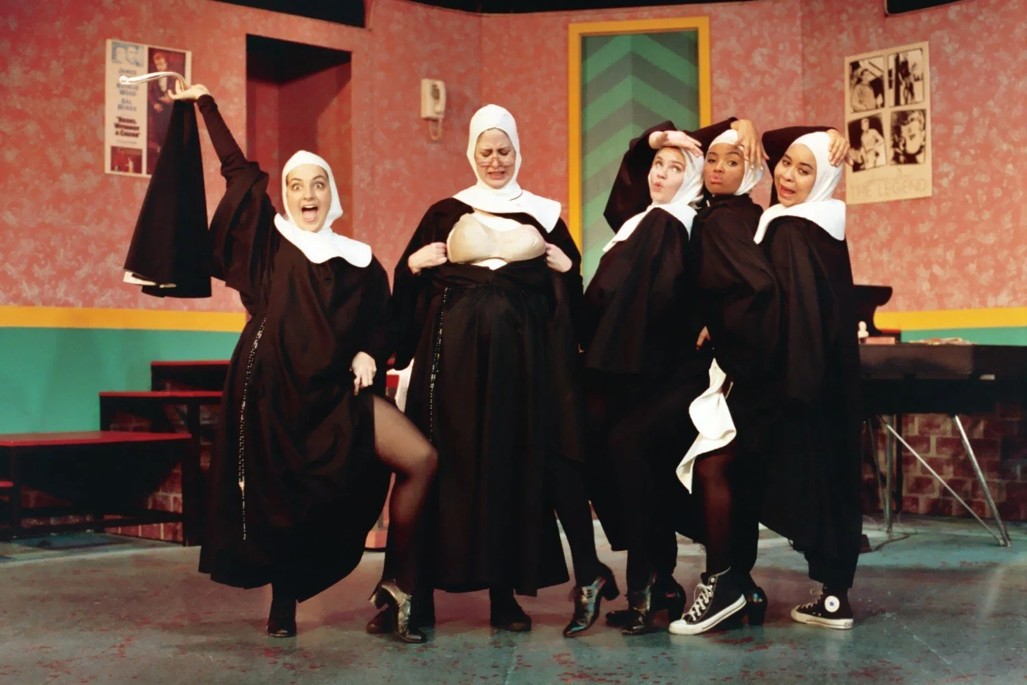 Five performers dressed as nuns, striking playful poses on stage in a colorful set.