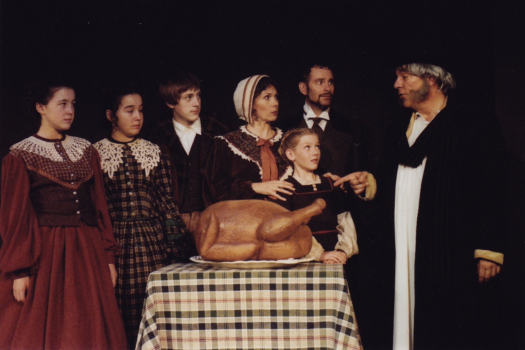 People in period costumes around a table with a roasted turkey on a checkered cloth, likely a scene from a theatrical production.