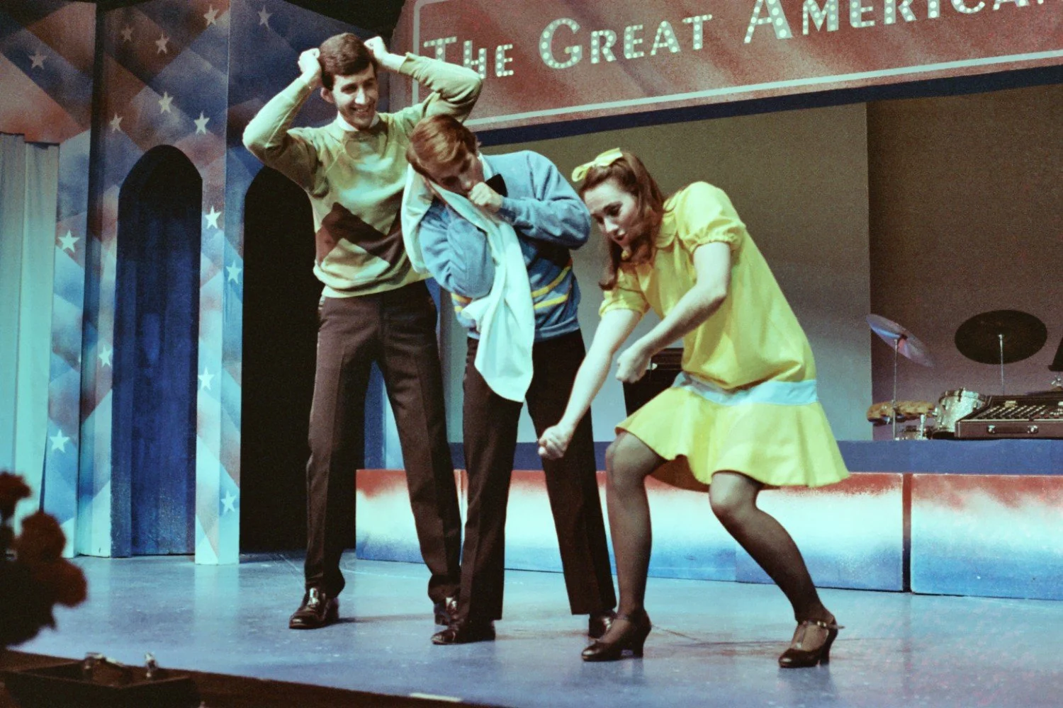 Three performers in vibrant 1960s-style costumes acting playfully on a stage with star-themed decorations and a sign reading 'The Great Americ...' in the background.