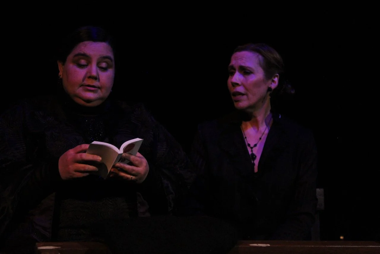 Two people in dark clothing in a theatrical setting, one holding a small book, the other looking at them.