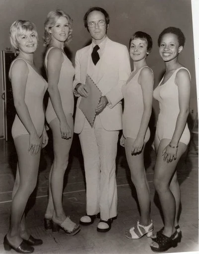 A man in a white suit holding a folder stands with four women in leotards and heels, posed for a photo indoors.