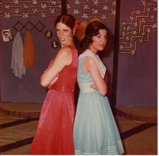 Two women wearing vintage dresses, one in red and the other in blue, posing back-to-back with arms crossed, in a theatrical setting featuring a patterned backdrop.