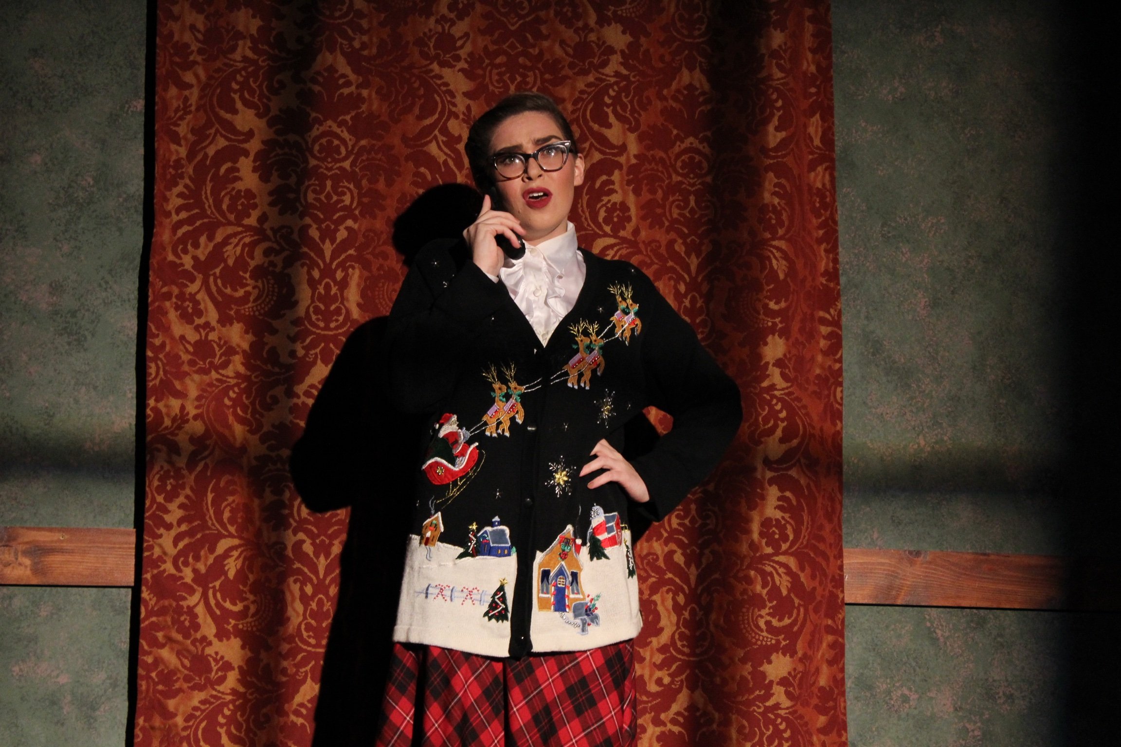 A person wearing a Christmas-themed sweater with reindeer and Santa Claus designs standing in front of a patterned curtain, holding a phone to their ear, with a plaid skirt and glasses.