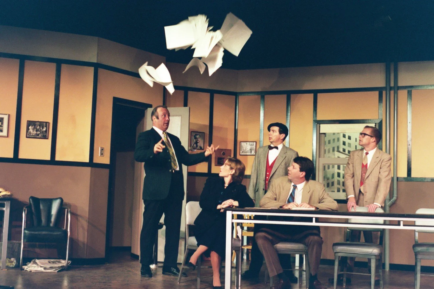 Stage drama scene with five actors in office setting, one standing and gesturing as papers fly overhead, others seated and reacting.