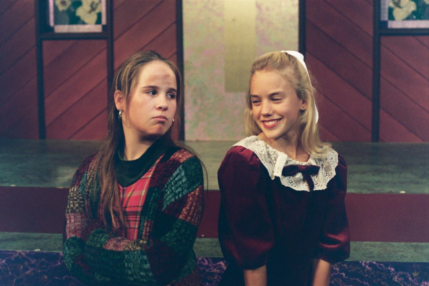 Two young girls in period-style clothing, one looking displeased, the other smiling, standing on a stage with a wooden backdrop.